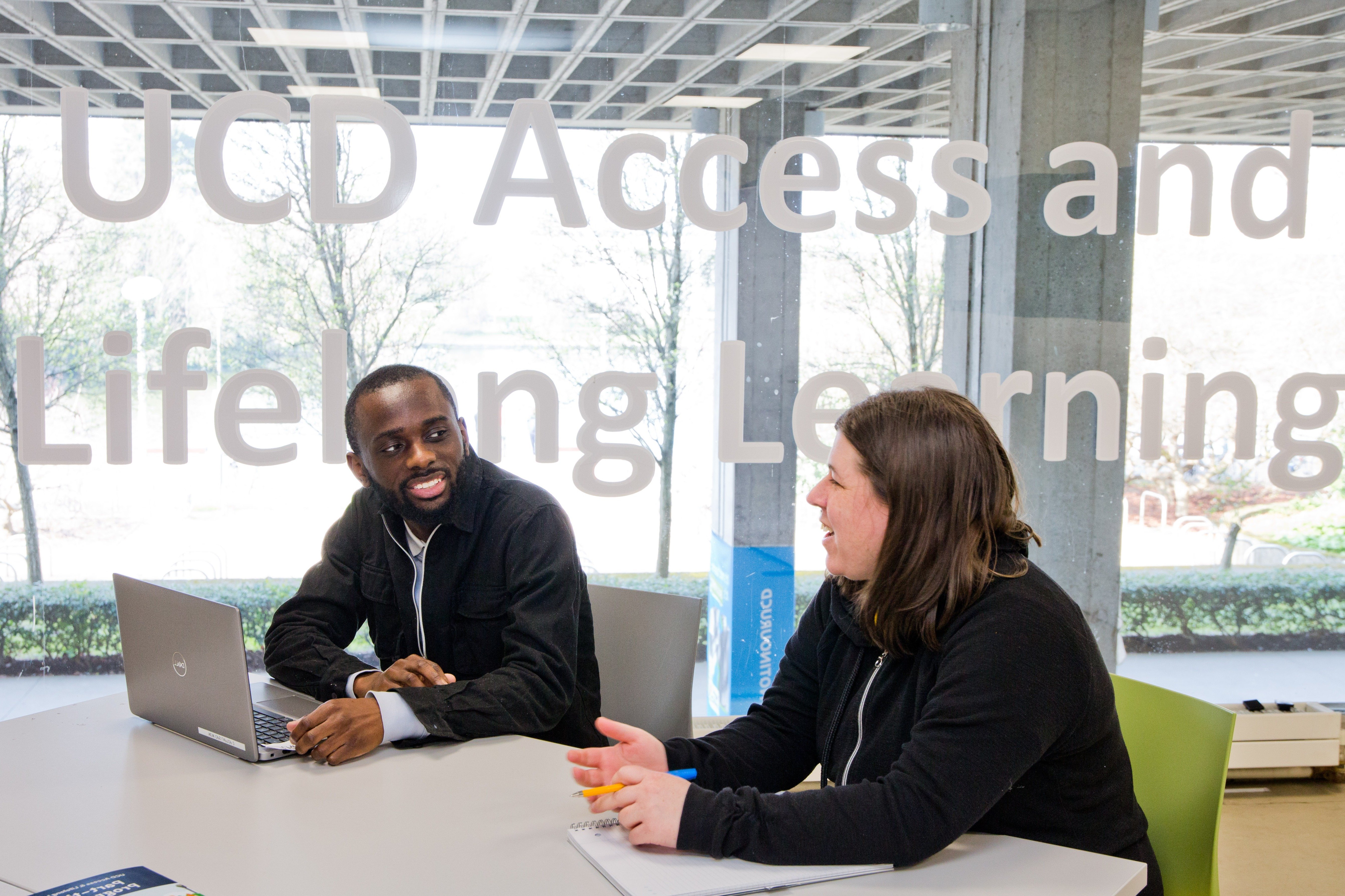 two persons sat at a desk and talking to each other in front of the Access and Lifelong Learning sign.