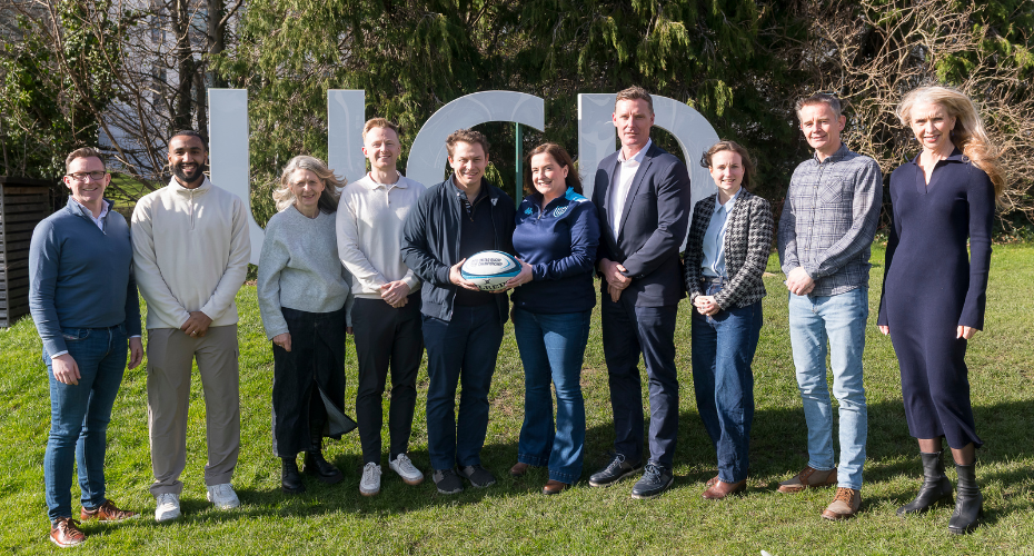 A group of people standing on grass in front of UCD sign and holding a rugby ball