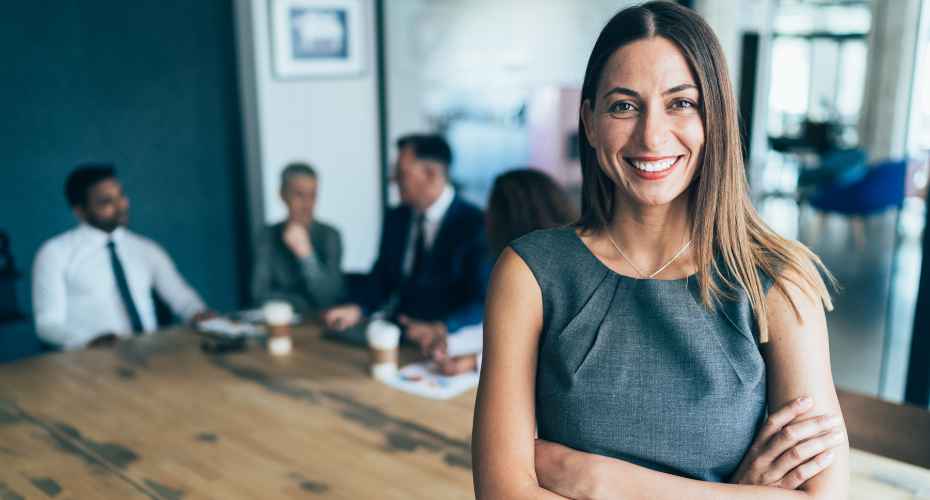 Business woman smiling to camera with her team slightly out of focus sitting at a table behind her