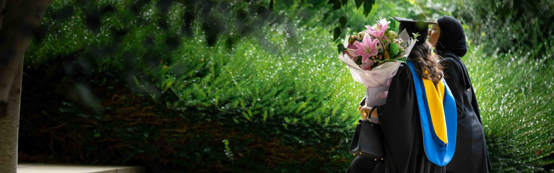 UCD graduate walking along a green path, holding a bouquet of flowers.