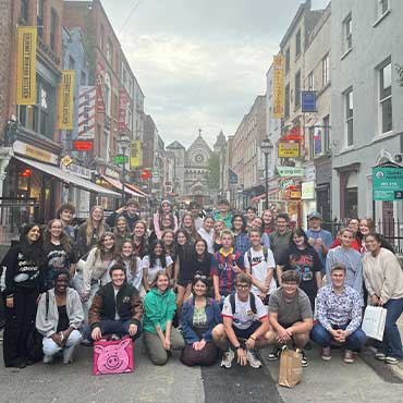 A large group of people pose together on a pedestrian street lined with shops and restaurants, with a church visible at the end of the street in the background.