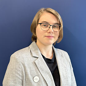 Young woman with glasses and short blonde hair, smiles to camera.