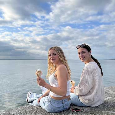 Two young women sit on a stone ledge by the water, eating ice cream and smiling at the camera under a partly cloudy sky.