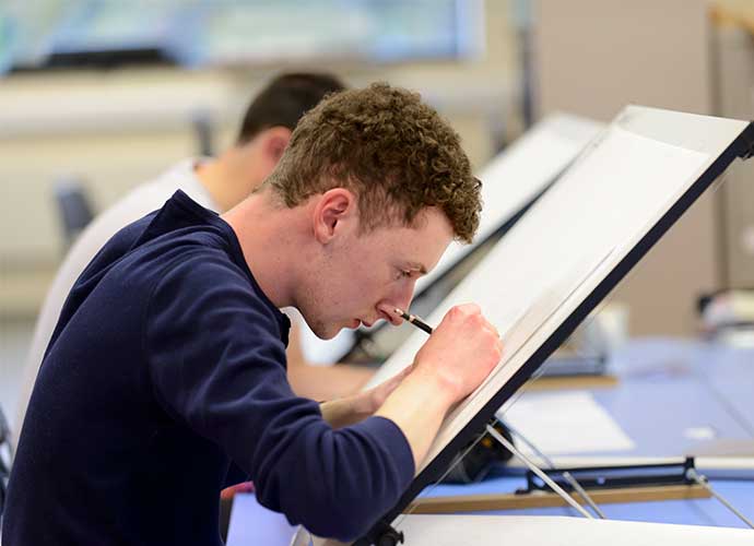 Young man concentrating while drawing on a drafting table with a pencil, in a classroom setting with another student working in the background.