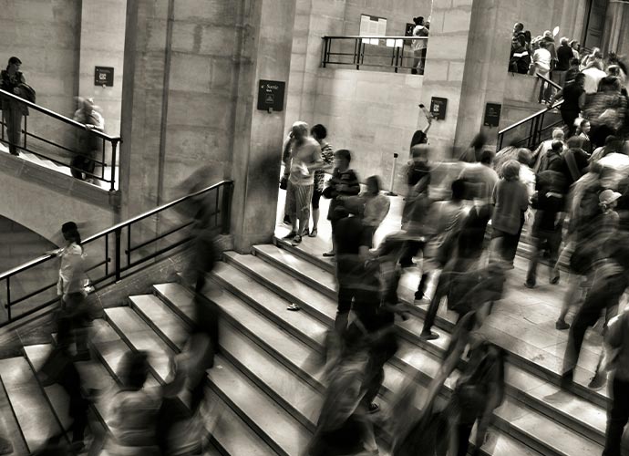 Black and white photo of a crowded staircase inside a large building, with many blurred figures moving up and down, suggesting hustle and bustle. A few people stand still near the walls while others hurry past.