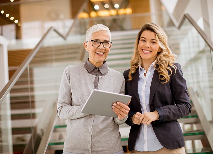 Two professional women standing indoors in front of a staircase, smiling at the camera. One has short gray hair, glasses, and is holding a tablet, while the other has long blonde hair and is wearing a dark blazer over a light shirt.