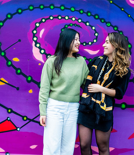 In front of a mural of a purple nebulous stand two young women.