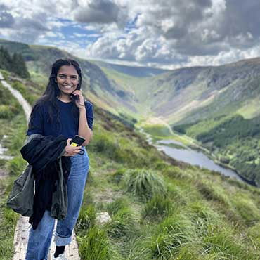 A student smiling while standing on a grassy trail overlooking a scenic valley with mountains, forest, and a lake under a partly cloudy sky.