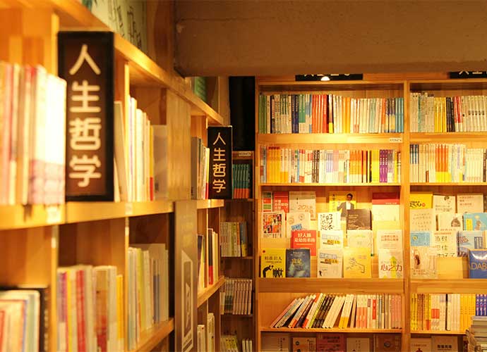 Bookshelves filled with colourful books in a warmly lit bookstore or library, with signs in Chinese characters displayed on the shelves.