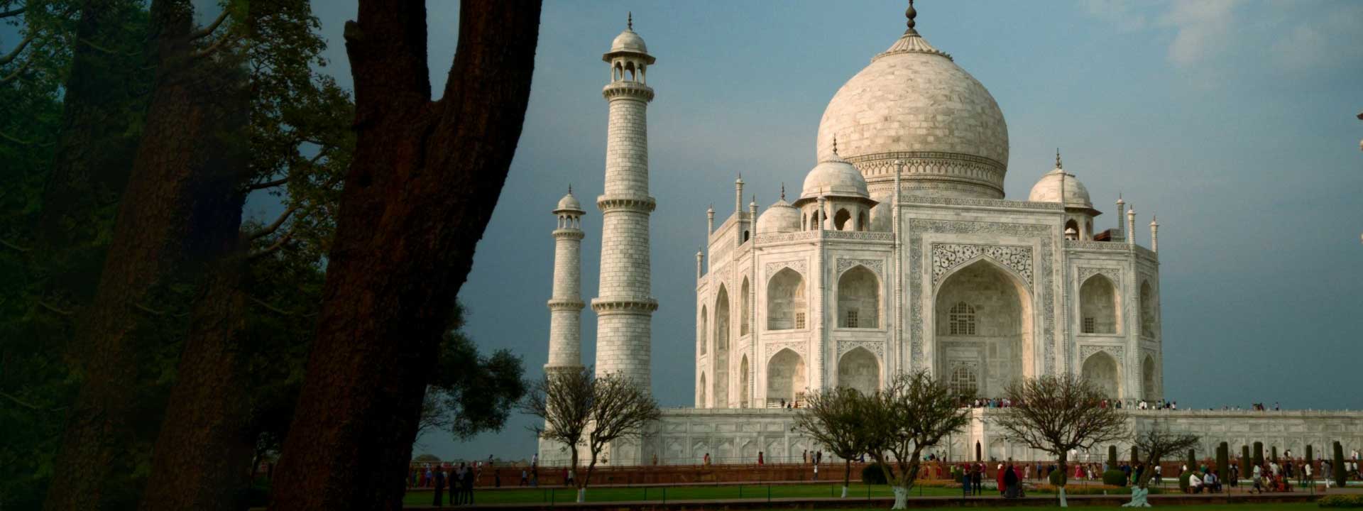 The Taj Mahal in Agra, India, with its white marble dome and minarets, set against a cloudy sky and framed by trees in the foreground, with visitors walking around the base.