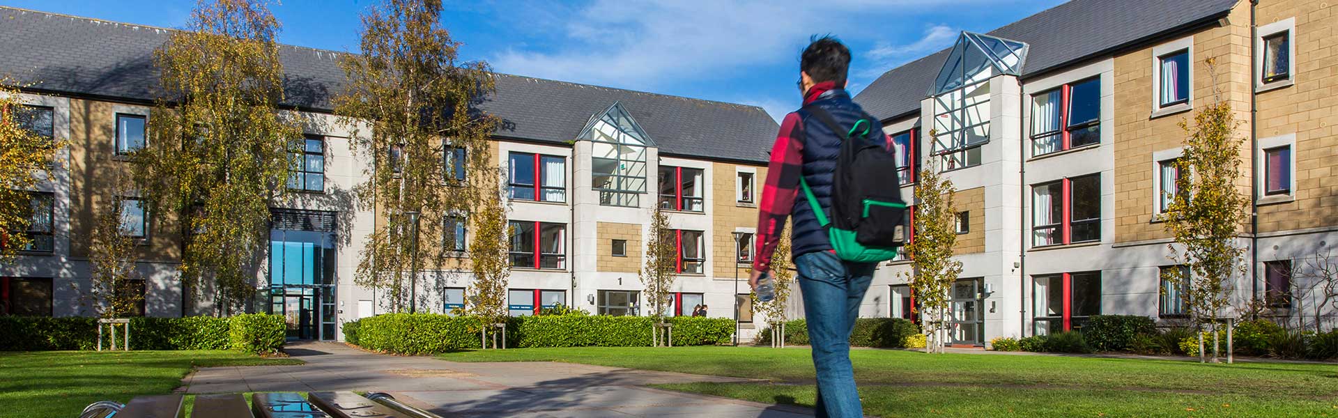 A student with a backpack walks toward a modern university accommodation building surrounded by trees and green lawns on a sunny day.