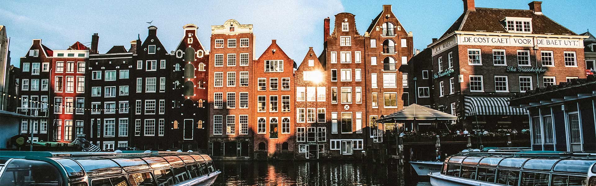 Row of narrow, gabled canal houses in Amsterdam, lit by evening sun, with tour boats docked on the canal in the foreground.