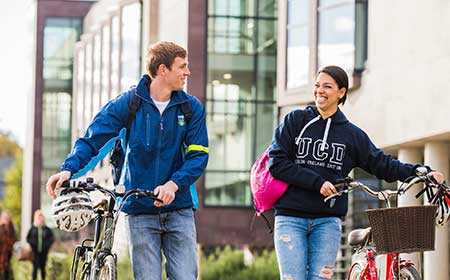 Two students walking with bicycles on campus, one wearing a UCD sweatshirt, outside a modern university building.