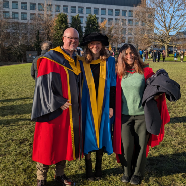 A UCD graduate and two UCD academics in caps and gowns outside on a sunny graduation day.