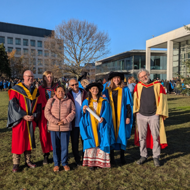 UCD graduates and UCD academic in caps and gowns outside on a sunny graduation day.