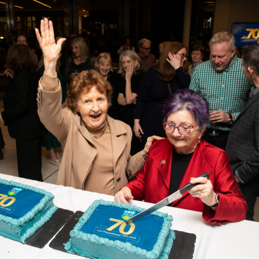 Woman cutting a cake surrounded by a group of people at the UCD Physiotherapy 70th anniversary celebration
