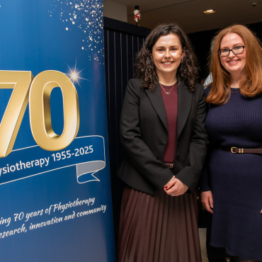 Two women standing beside a UCD Physiotherapy 70th anniversary celebration banner