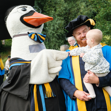 PhD graduate holding a baby shaking hands with UCD swan mascot