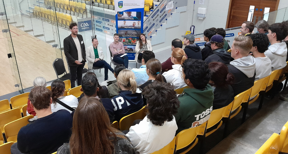 A man standing beside three seated people presenting to a room full of people in a sports hall